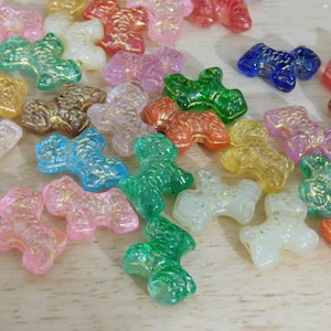 May include: A collection of colorful, translucent dog-shaped beads in various colors, including pink, green, blue, and gold. The beads have a glittery, textured appearance and are scattered on a light-colored surface.