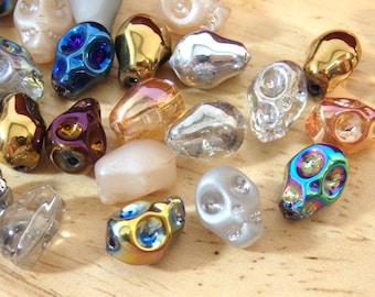 Könnte beinhalten: Eine Sammlung bunter Glasschädelperlen in verschiedenen Formen und Größen. Die Perlen sind irisierend und haben verschiedene Oberflächen, darunter klar, matt und metallisch.