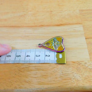 May include: A yellow and iridescent metal charm shaped like a heart with a Ouija board design and a butterfly inside. The charm is 5 cm long and is measured against a ruler.
