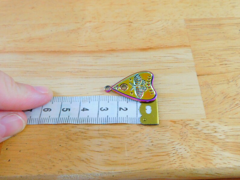 May include: A yellow and iridescent metal charm shaped like a heart with a Ouija board design and a butterfly inside. The charm is 5 cm long and is measured against a ruler.