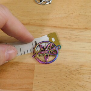 K&ouml;nnte beinhalten: Ein regenbogenfarbener, metallischer Anh&auml;nger mit einem Pentagramm und einem Drachenmotiv. Der Anh&auml;nger wird mit einem Lineal gemessen.