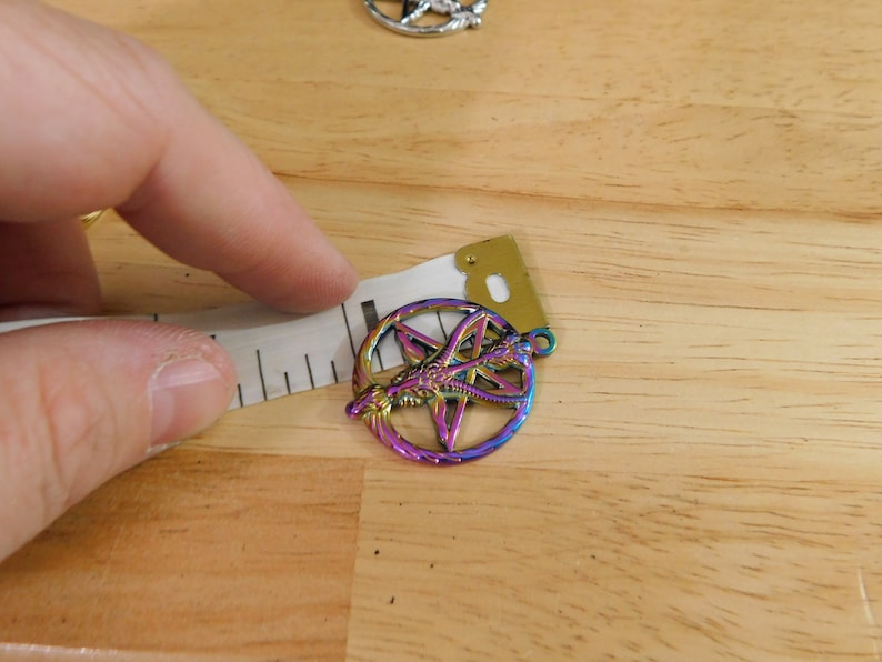 K&ouml;nnte beinhalten: Ein regenbogenfarbener, metallischer Anh&auml;nger mit einem Pentagramm und einem Drachenmotiv. Der Anh&auml;nger wird mit einem Lineal gemessen.