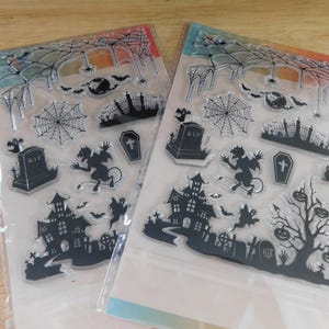 May include: Two clear Halloween-themed stamp sets. Each set features black illustrations of haunted houses, tombstones, bats, cobwebs, and a devilish figure. The sets are packaged on a white backing with colorful accents.