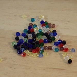 May include: A collection of colorful glass beads in various shades of blue, green, red, yellow, and black. The beads are scattered on a light wood surface.