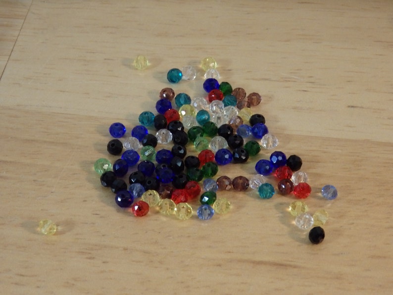 May include: A collection of colorful glass beads in various shades of blue, green, red, yellow, and black. The beads are scattered on a light wood surface.