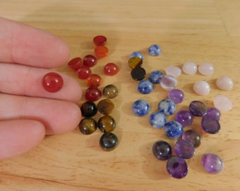 May include: Assortment of round cabochon gemstones in various colors, including red, orange, brown, blue, purple, and pink. The stones are arranged in rows on a wooden surface.