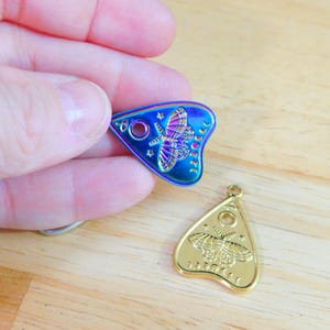 May include: Two gold-toned and iridescent charm pendants in the shape of a triangle with a moth design and the words "Ouija" and "Planchette".