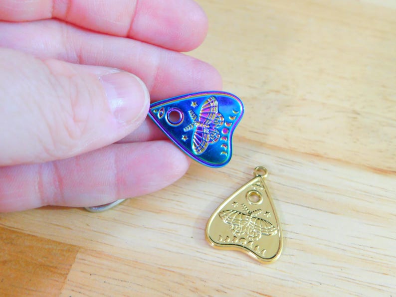 May include: Two gold-toned and iridescent charm pendants in the shape of a triangle with a moth design and the words "Ouija" and "Planchette".