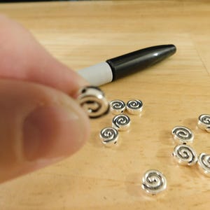 May include: A close-up of silver metal beads with a spiral design. The beads are scattered on a wooden surface.