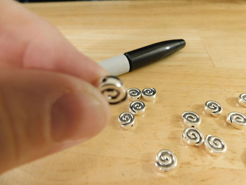 May include: A close-up of silver metal beads with a spiral design. The beads are scattered on a wooden surface.