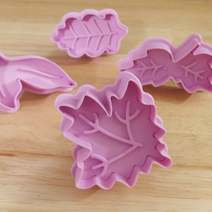 May include: Four pink plastic leaf-shaped cookie cutters. The cutters are in various leaf designs, including maple and oak leaves. The cutters are on a light brown wooden surface.
