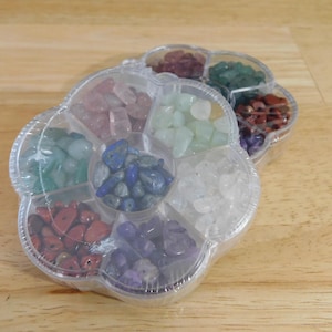 May include: A clear, flower-shaped container filled with various colorful gemstones. The gemstones are in shades of red, green, blue, purple, pink, and white. The container is sitting on a light brown wooden surface.