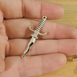 May include: Silver-toned metal sword charm with a detailed design. The charm has a pointed tip and a cross guard.
