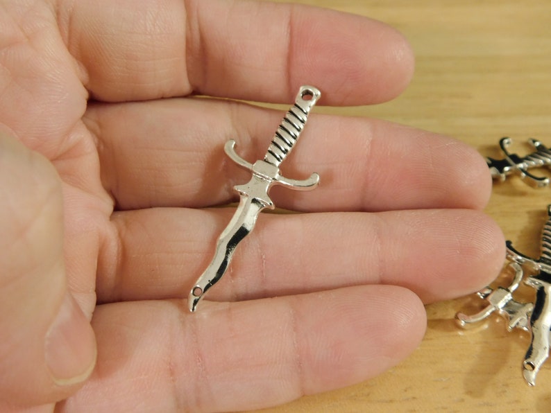 May include: Silver-toned metal sword charm with a detailed design. The charm has a pointed tip and a cross guard.