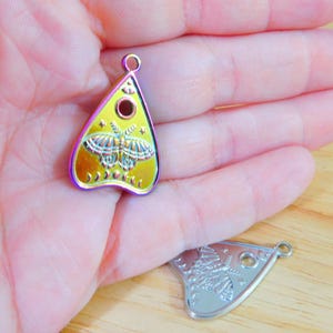 May include: A gold-coloured metal charm shaped like a witch's board or planchette with a butterfly design and a black circle in the centre. The charm has a rainbow iridescent finish.