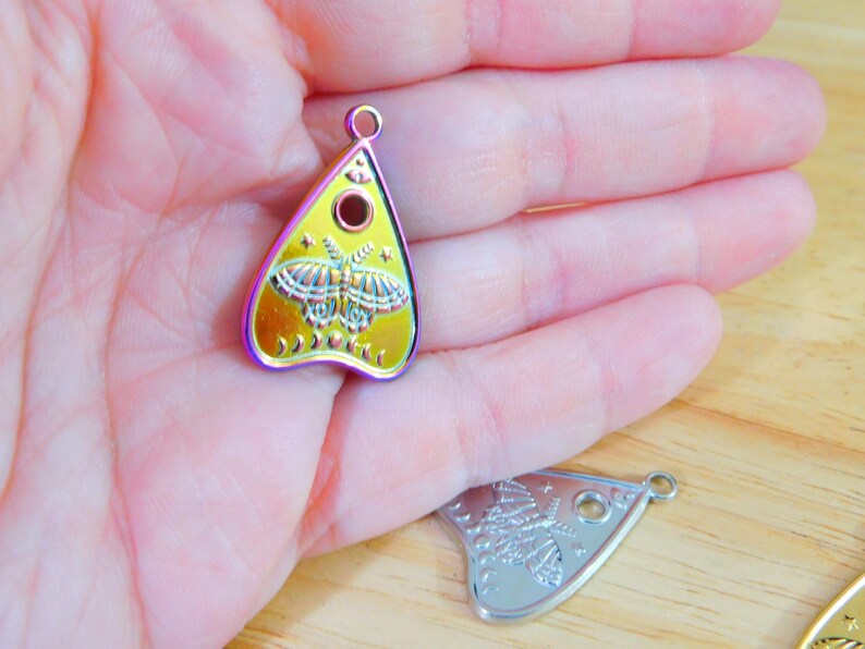 May include: A gold-coloured metal charm shaped like a witch's board or planchette with a butterfly design and a black circle in the centre. The charm has a rainbow iridescent finish.