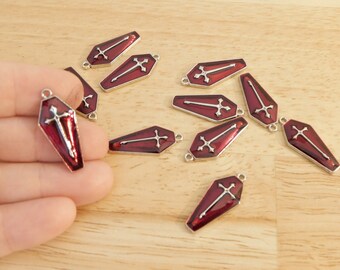 May include: Ten red metal coffin charms with silver crosses. The charms have a loop at the top for attaching to jewelry or other crafts.