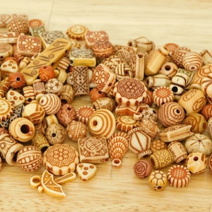 May include: A collection of wooden beads with various patterns and designs. The beads are in shades of brown and beige, and some have a reddish-brown hue. The beads are arranged in a pile on a light brown wooden surface.