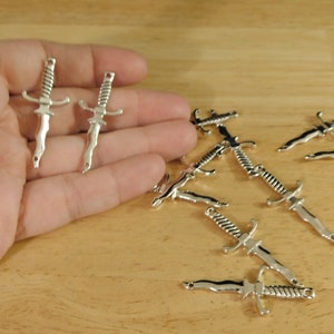 May include: A collection of silver-toned sword charms. The charms are detailed with a twisted handle and a pointed tip. The charms are scattered on a wooden surface.