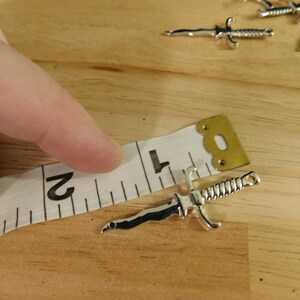 May include: A silver-toned dagger charm measuring 2 inches in length. The charm is lying on a white measuring tape with black markings.