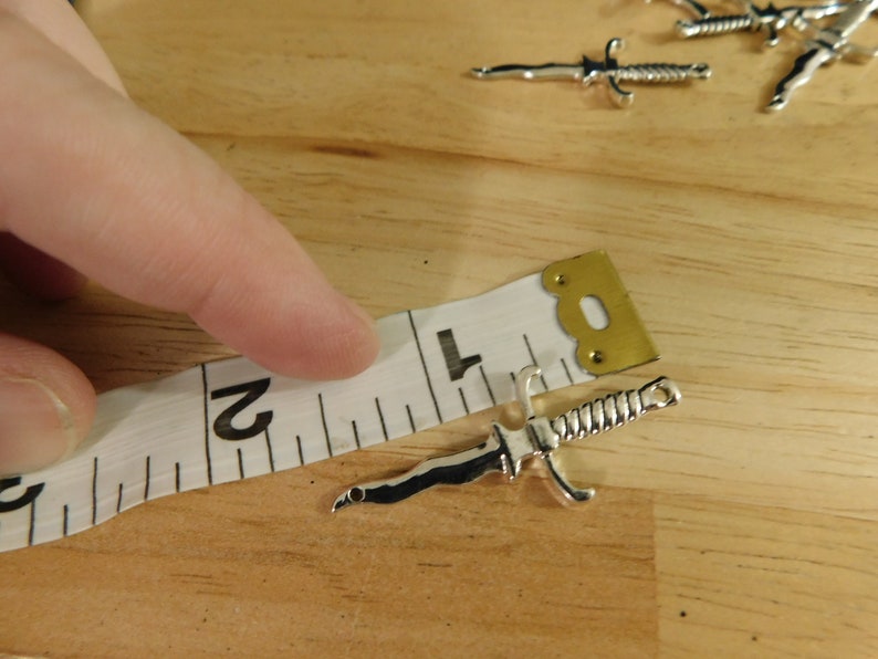 May include: A silver-toned dagger charm measuring 2 inches in length. The charm is lying on a white measuring tape with black markings.