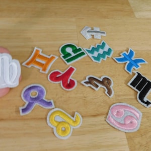May include: A collection of colorful embroidered zodiac patches, including Virgo, Libra, and Cancer, with white borders. The patches are in various colors, such as orange, green, and blue, and are arranged on a wooden surface.