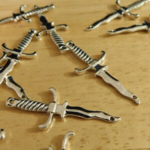 May include: A group of silver-toned dagger charms with intricate details. The charms are scattered on a wooden surface.