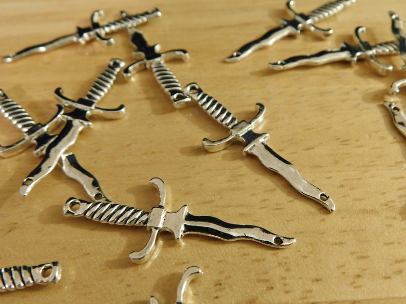 May include: A group of silver-toned dagger charms with intricate details. The charms are scattered on a wooden surface.