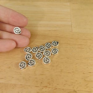 May include: A group of silver metal beads with a spiral design. The beads are arranged in a loose pile on a wooden surface.