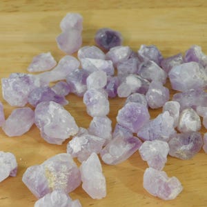 May include: A collection of raw amethyst crystals in various sizes and shapes. The crystals display a range of purple hues, from pale lavender to deep violet, and are scattered on a light brown wooden surface.