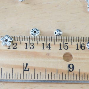 May include: Close-up of various silver-colored beads, including flower-shaped and cylindrical designs. A wooden ruler with inch markings is in the foreground, with beads scattered around. The beads are likely for jewelry making.