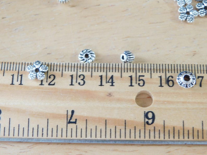 May include: Close-up of various silver-colored beads, including flower-shaped and cylindrical designs. A wooden ruler with inch markings is in the foreground, with beads scattered around. The beads are likely for jewelry making.