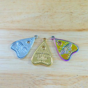 May include: Three metal charms in the shape of a witch's board planchette. The charms are silver, gold, and iridescent.