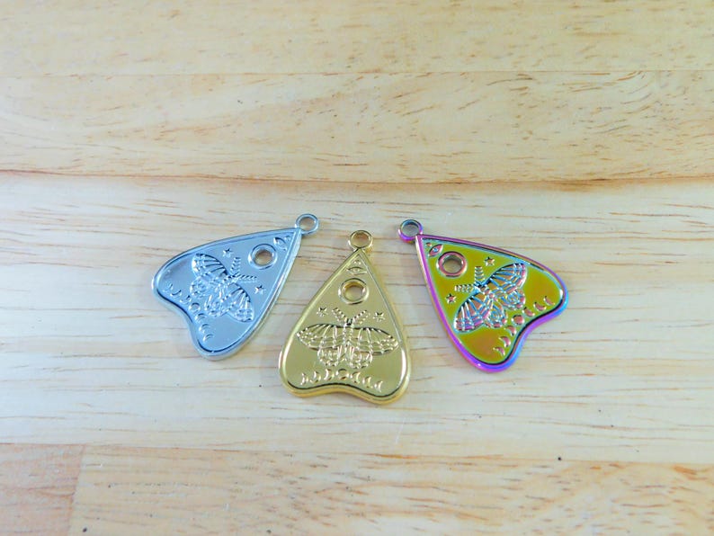 May include: Three metal charms in the shape of a witch's board planchette. The charms are silver, gold, and iridescent.