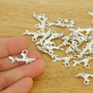 May include: A pile of silver shark charm pendants with a single shark charm pendant held in a hand. The charms are about 1 inch long and have a loop at the top for attaching to jewelry.