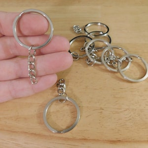 May include: A collection of silver-colored key rings with attached chains. Several rings are scattered on a light brown wooden surface. One ring is held in a hand, showcasing the chain and ring design. The rings are made of metal and are suitable for keychains.