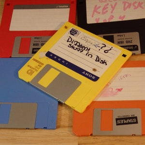 May include: A collection of five floppy disks, including a yellow Sony disk labeled "DiAnent Stup? in Disk" and a blue disk labeled "HD Fujifilm 3.5" with a Staples label on the orange disk.