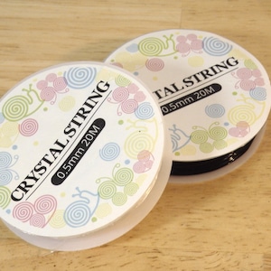 May include: Two spools of black elastic string, each with a white label that says "CRYSTAL STRING 0.5mm 20M" and a colorful design of swirls and butterflies.