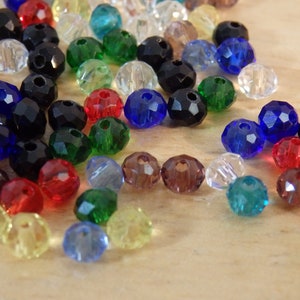 May include: Assortment of colorful faceted glass beads in various colors, including black, blue, green, red, clear, and yellow. The beads are scattered on a wooden surface.