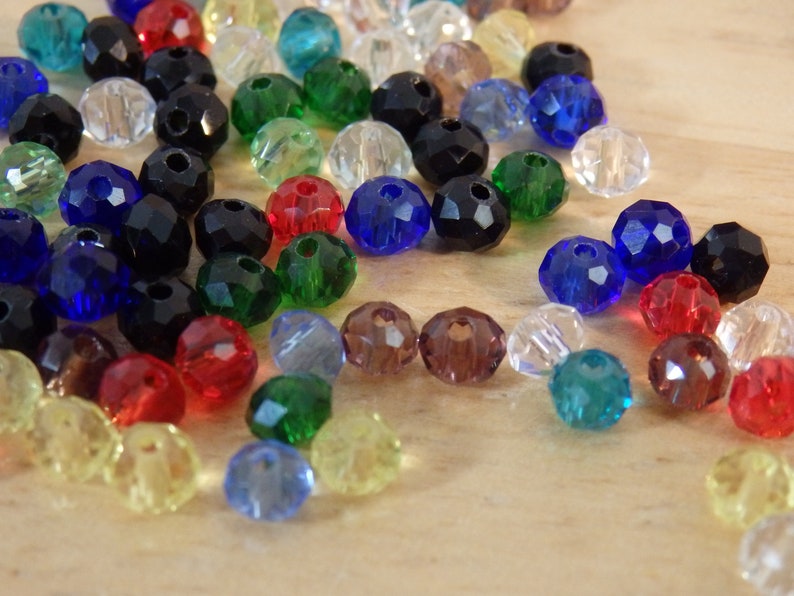 May include: Assortment of colorful faceted glass beads in various colors, including black, blue, green, red, clear, and yellow. The beads are scattered on a wooden surface.