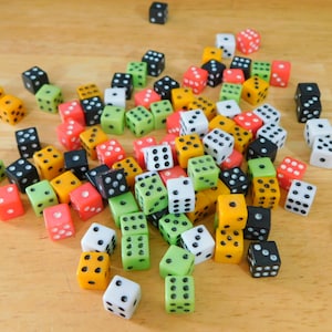 May include: A collection of colorful dice scattered on a light brown wooden surface. The dice are in various colors, including red, green, yellow, black, and white, with black dots indicating the numbers. A game accessory.