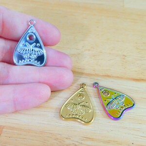 May include: Three silver, gold, and iridescent charm pendants in the shape of a witch's board planchette with a moth design. The planchette has a hole in the centre and the words "Planchette" written on the bottom.