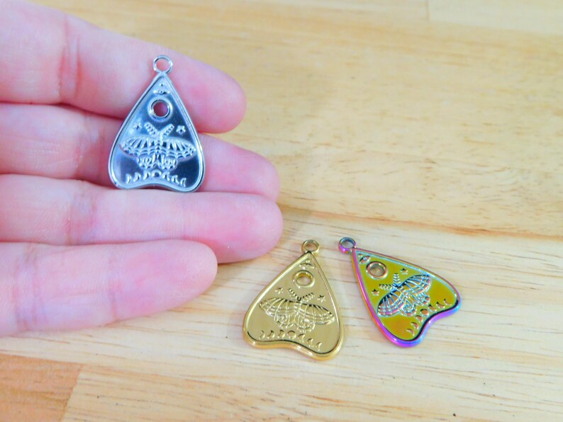 May include: Three silver, gold, and iridescent charm pendants in the shape of a witch's board planchette with a moth design. The planchette has a hole in the centre and the words "Planchette" written on the bottom.