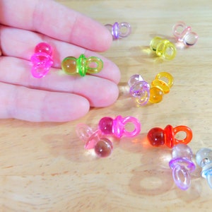 May include: Assortment of colorful plastic baby pacifier charms. The charms are translucent and come in pink, green, yellow, orange, red, and purple.