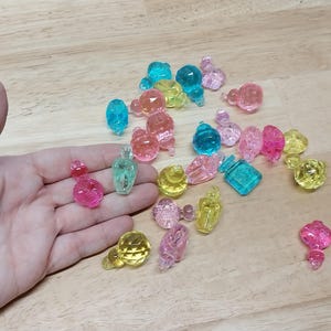 May include: A collection of small, translucent, multi-colored plastic beads shaped like miniature perfume bottles or decorative ornaments. The beads feature faceted surfaces in shades of pink, yellow, blue, and clear, with some pieces showing a distinct bottle-like silhouette and others appearing as rounded or geometric shapes.