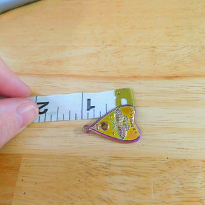 May include: A yellow, iridescent heart-shaped charm with a black and white image of a moth and the text "Ouija" on it. The charm is being measured with a tape measure.