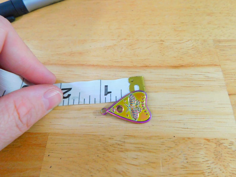 May include: A yellow, iridescent heart-shaped charm with a black and white image of a moth and the text "Ouija" on it. The charm is being measured with a tape measure.