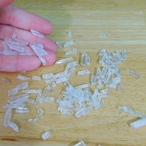 May include: A collection of small, clear quartz crystals scattered on a wooden surface. The crystals are various shapes and sizes, some are long and thin, others are shorter and thicker.