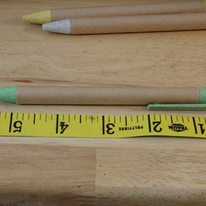 Op de afbeelding: Een groene en bruine pen met een bruin papieren lichaam en een groene plastic punt en dop. De pen ligt op een gele meetlint met zwarte markeringen. De tape leest "1, 2, 3, 4, 5, 6, 7, 8, 9" in inches.