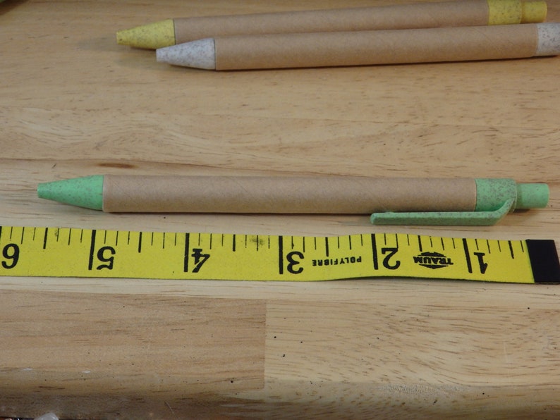 Op de afbeelding: Een groene en bruine pen met een bruin papieren lichaam en een groene plastic punt en dop. De pen ligt op een gele meetlint met zwarte markeringen. De tape leest "1, 2, 3, 4, 5, 6, 7, 8, 9" in inches.